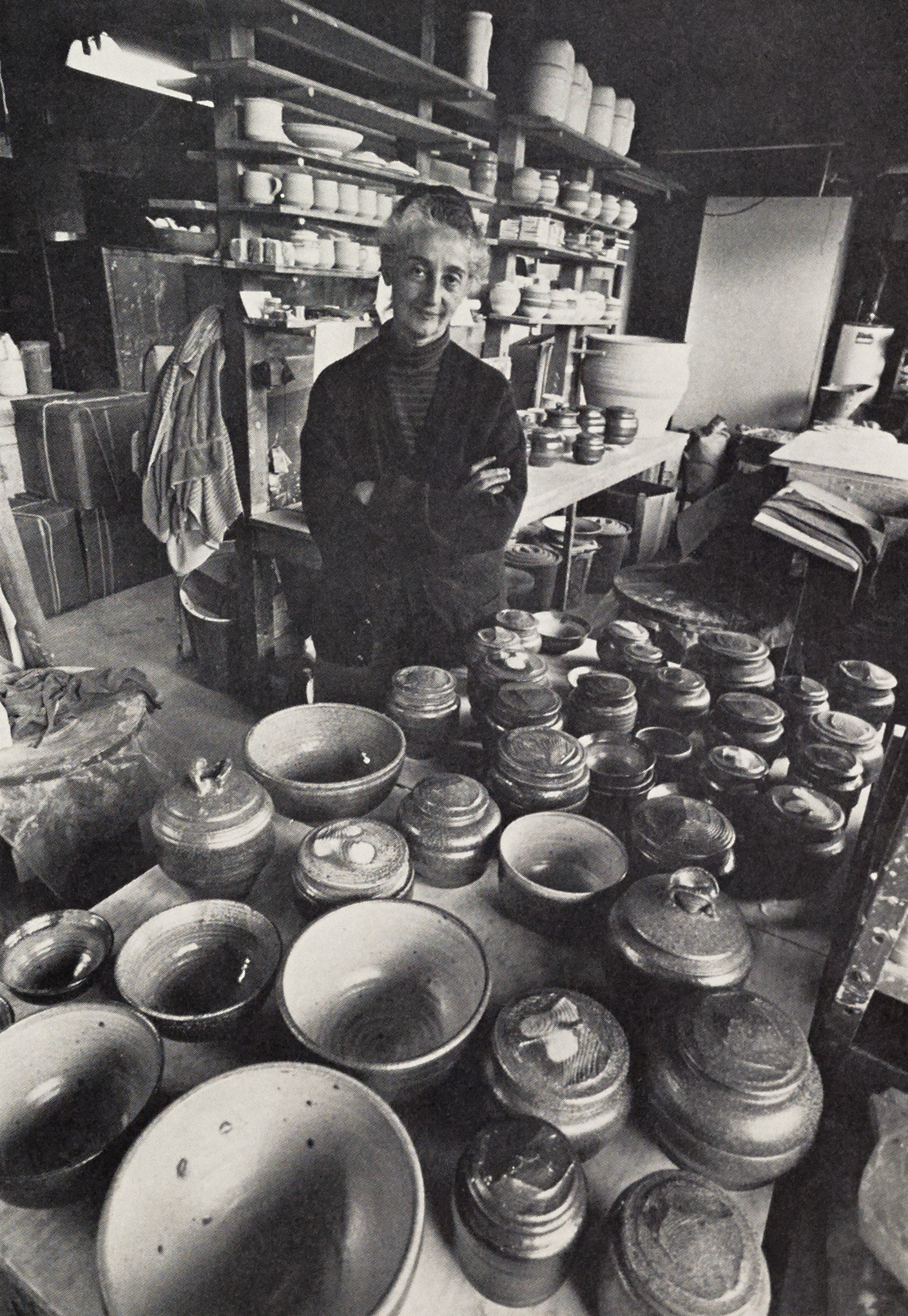 Black and white photograph in pottery studio with giant table filled with various shaped ceramic bowls and lidded pots. Standing behind is an older woman with arms crossed wearing a black sweater and dark turtleneck. Behind her are shelves of more light colored ceramic pieces and equipment.