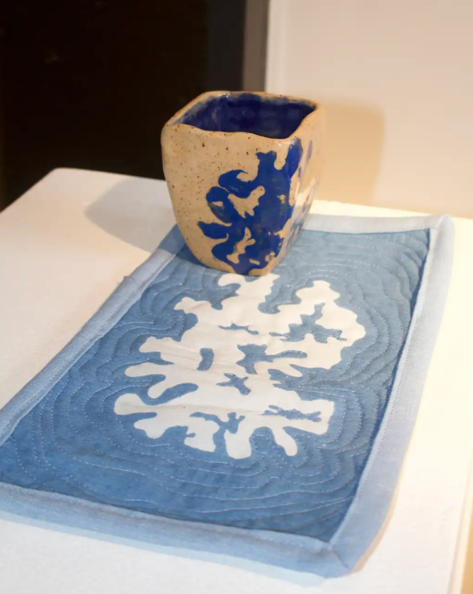 Same Story: A Conversation in Clay and Cloth. This indigo-inspired installation brings together clay, cloth, and driftwood – each carrying the same hand-cut pattern, a visual echo between mediums. The design moves from dyed fabric to ceramic form, uniting creative practices into one shared story. Here, material becomes language; touch becomes translation; and clay and cloth speak, at last, in the same voice.